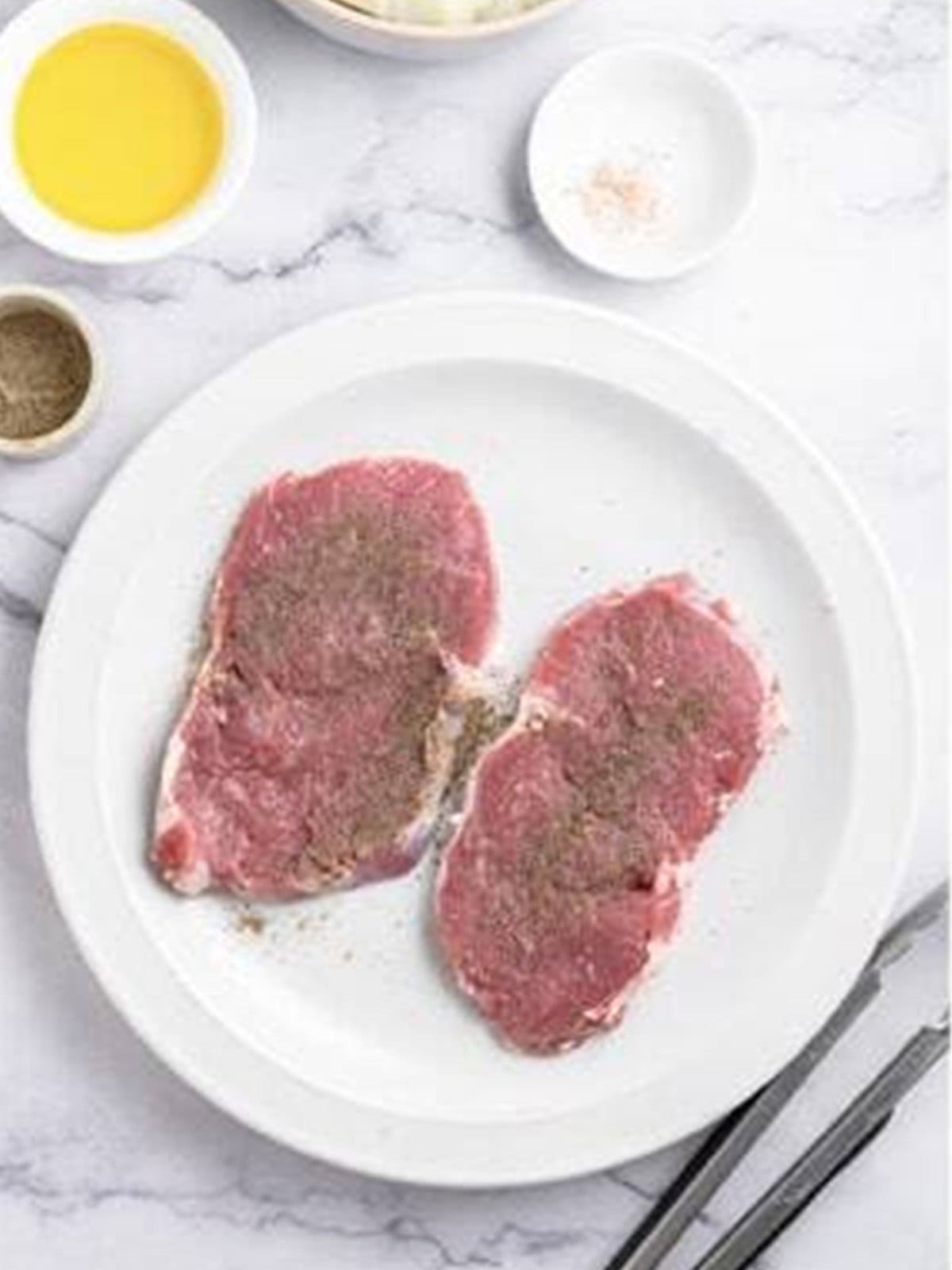 Thin ribeye steaks seasoned with salt and pepper on a white plate.