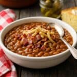 A white bowl of homemade stove top baked beans topped with crispy bacon and golden french fried onions on a rustic table.