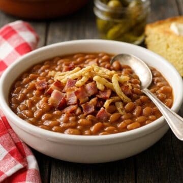 A white bowl of homemade stove top baked beans topped with crispy bacon and golden french fried onions on a rustic table.