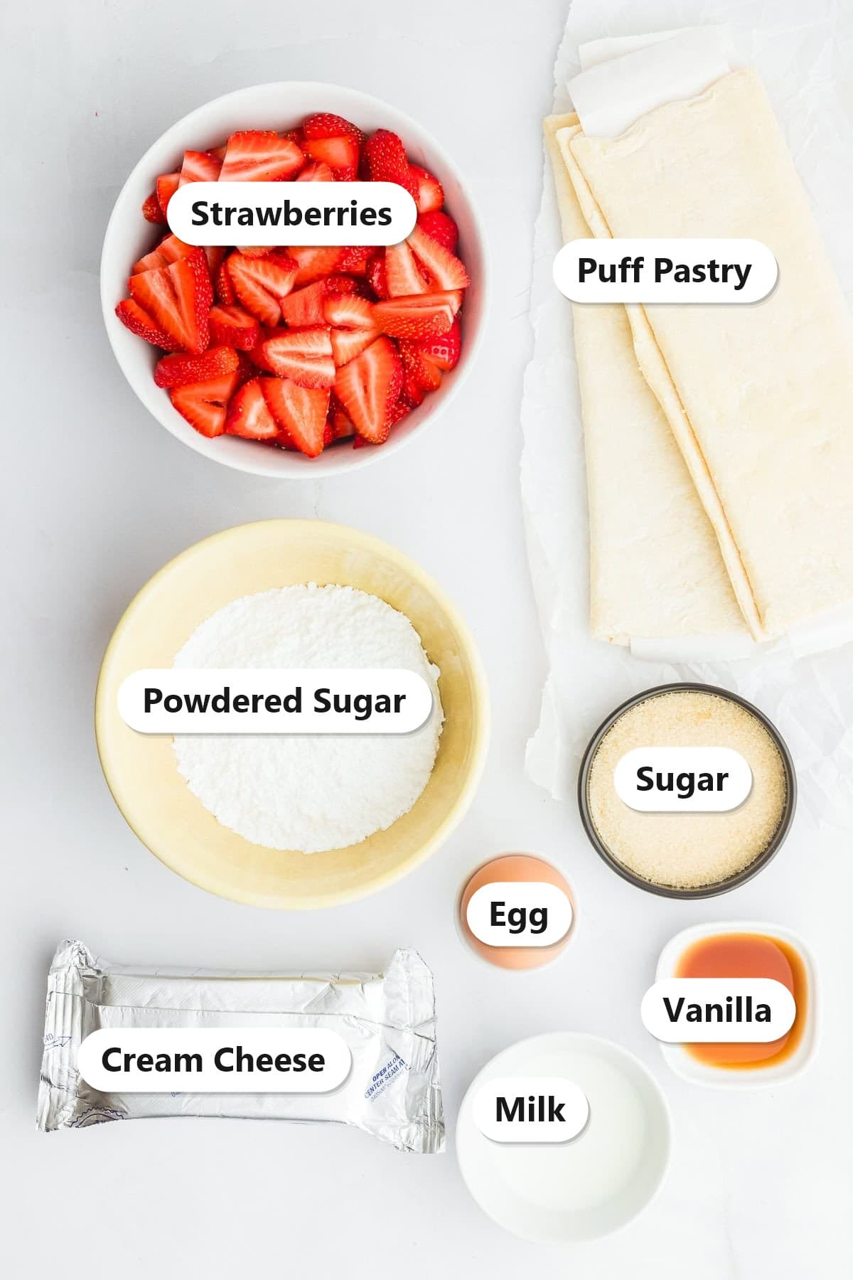 Ingredients to make strawberry cream cheese danishes labeled including fresh strawberries, puff pastry, powdered sugar, sugar, egg, vanilla, cream cheese, and milk.