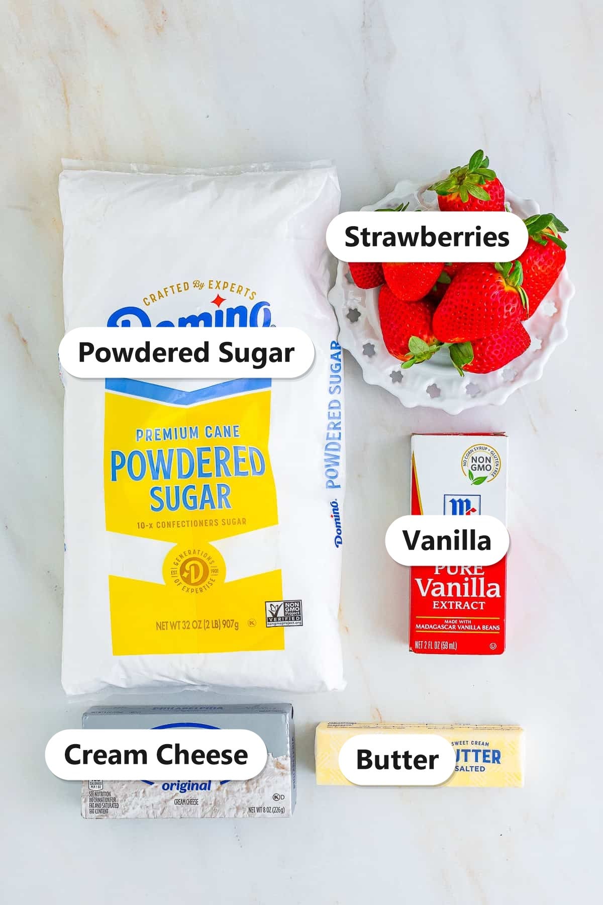 Strawberry cream cheese frosting ingredients including fresh strawberries, cream cheese, butter, vanilla, and powdered sugar labeled on a marble surface.