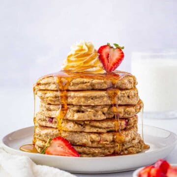 A stack of strawberry oatmeal pancakes drizzled with maple syrup and topped with fresh strawberries.