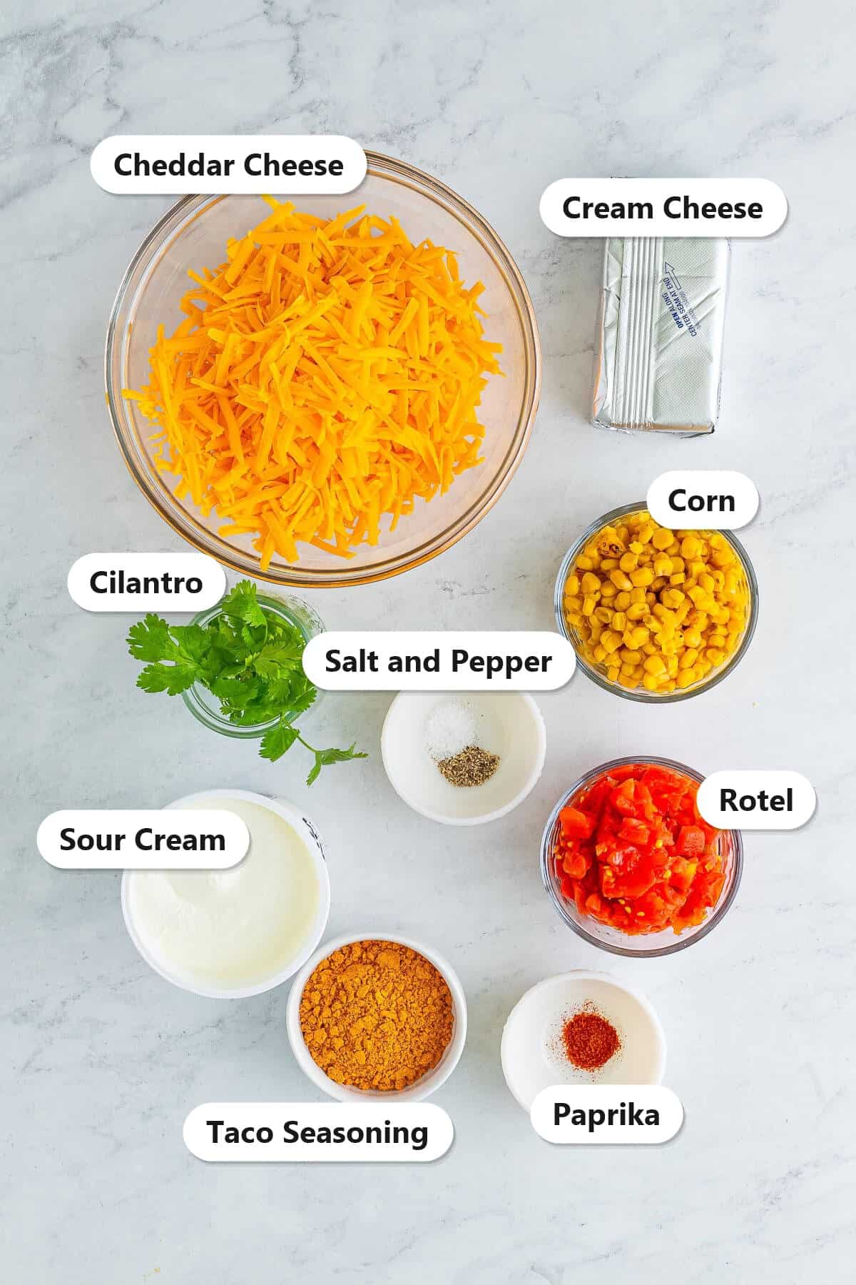 Labeled ingredients for taco dip including cheddar cheese, cream cheese, sour cream, corn, Rotel, taco seasoning, cilantro, salt, pepper, and paprika.