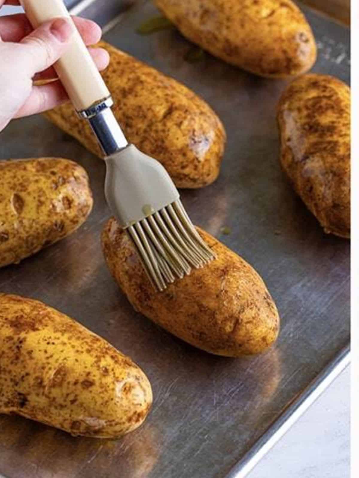 Russet potatoes being brushed with olive oil on a baking tray.