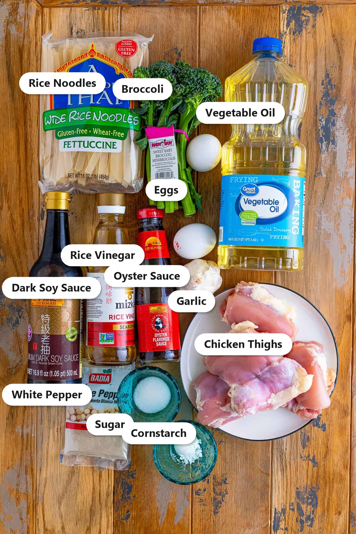 Pad see ew ingredients including wide rice noodles, chicken thighs, broccolini, eggs, dark soy sauce, oyster sauce, rice vinegar, garlic, and sugar labeled on a wood surface.