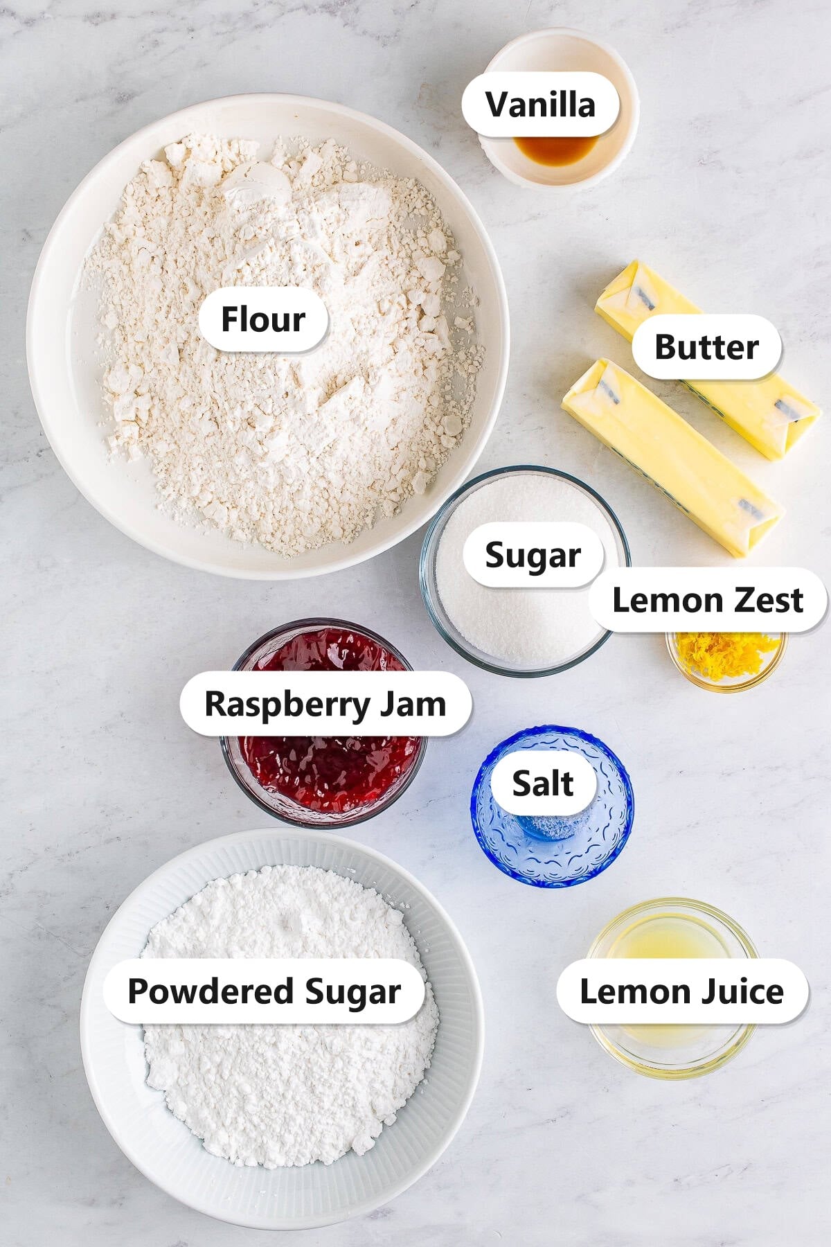 Labeled ingredients to make thumbprint cookies including flour, butter, sugar, powdered sugar, vanilla, salt, raspberry jam, lemon zest, and lemon juice.