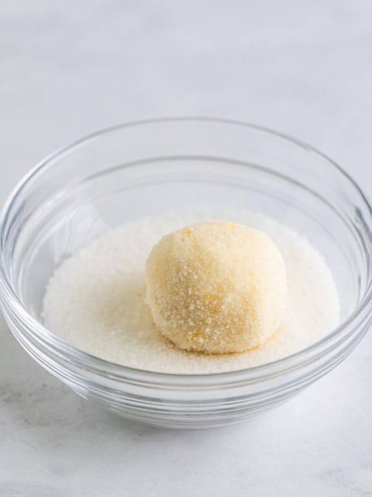 A thumbprint cookie dough ball rolled in granulated sugar in a glass bowl.