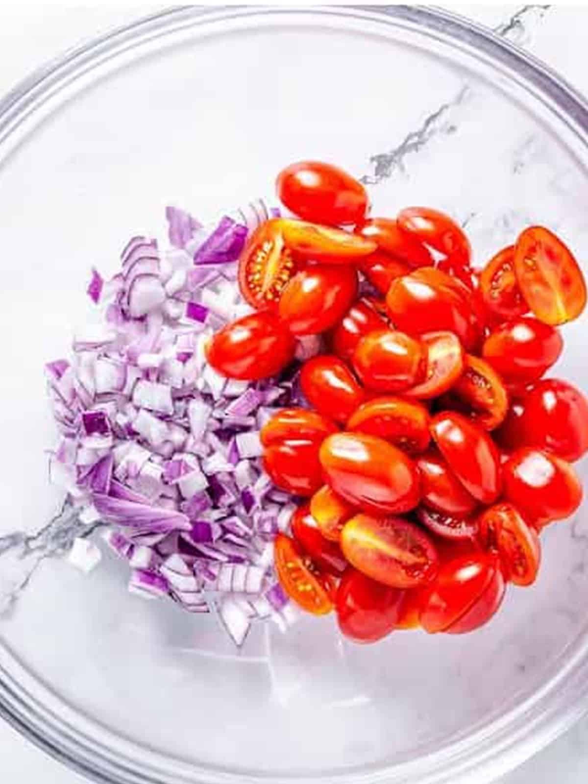Halved grape tomatoes and diced red onion together in a glass mixing bowl.