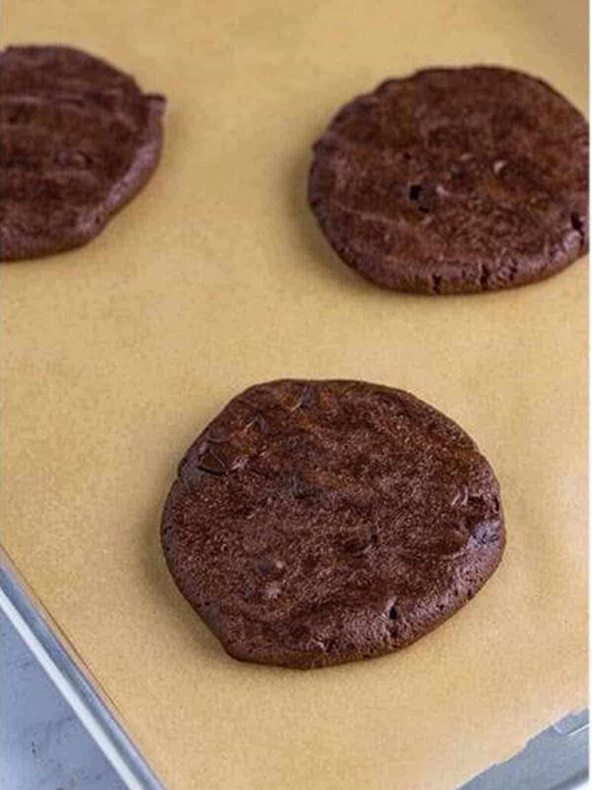 Large triple chocolate cookie dough balls pressed flat on a parchment lined sheet tray.