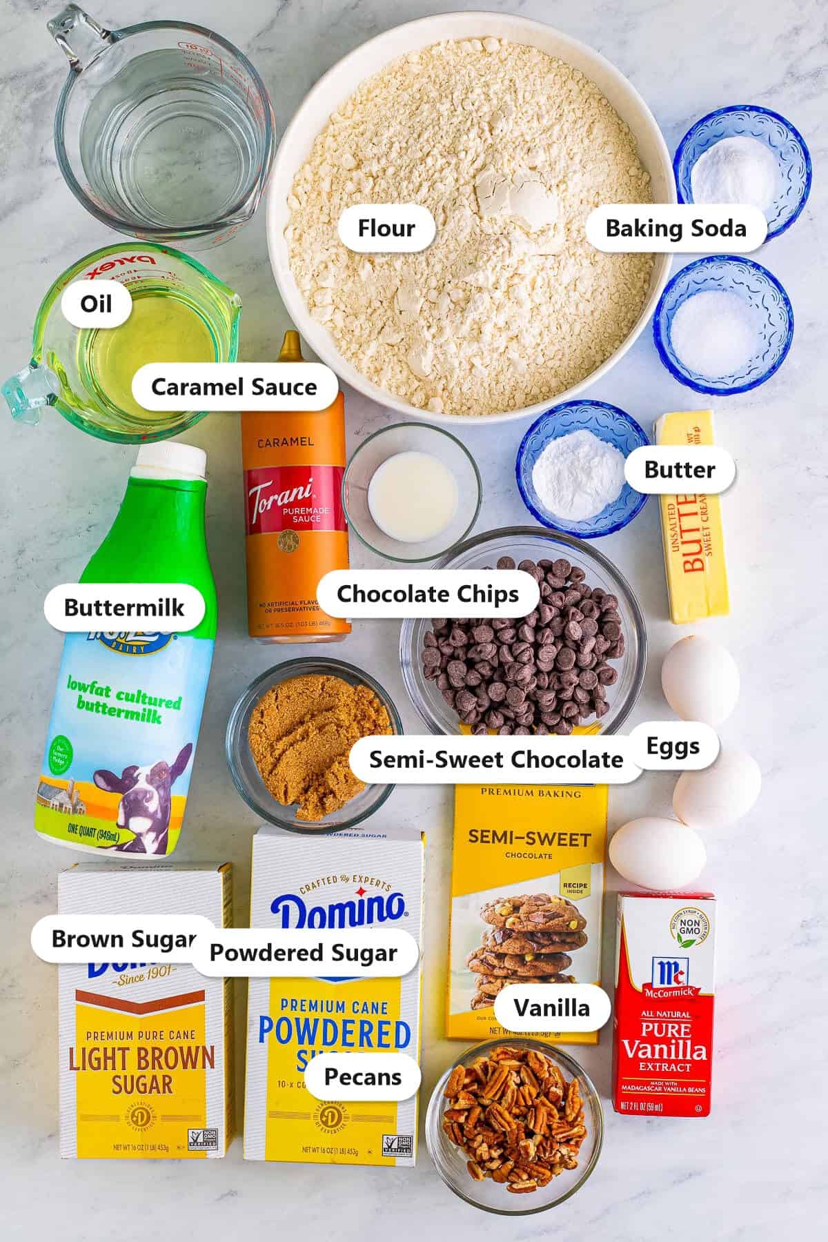 Labeled ingredients for chocolate turtle cake including flour, buttermilk, oil, eggs, chocolate, brown sugar, powdered sugar, caramel sauce, and pecans.