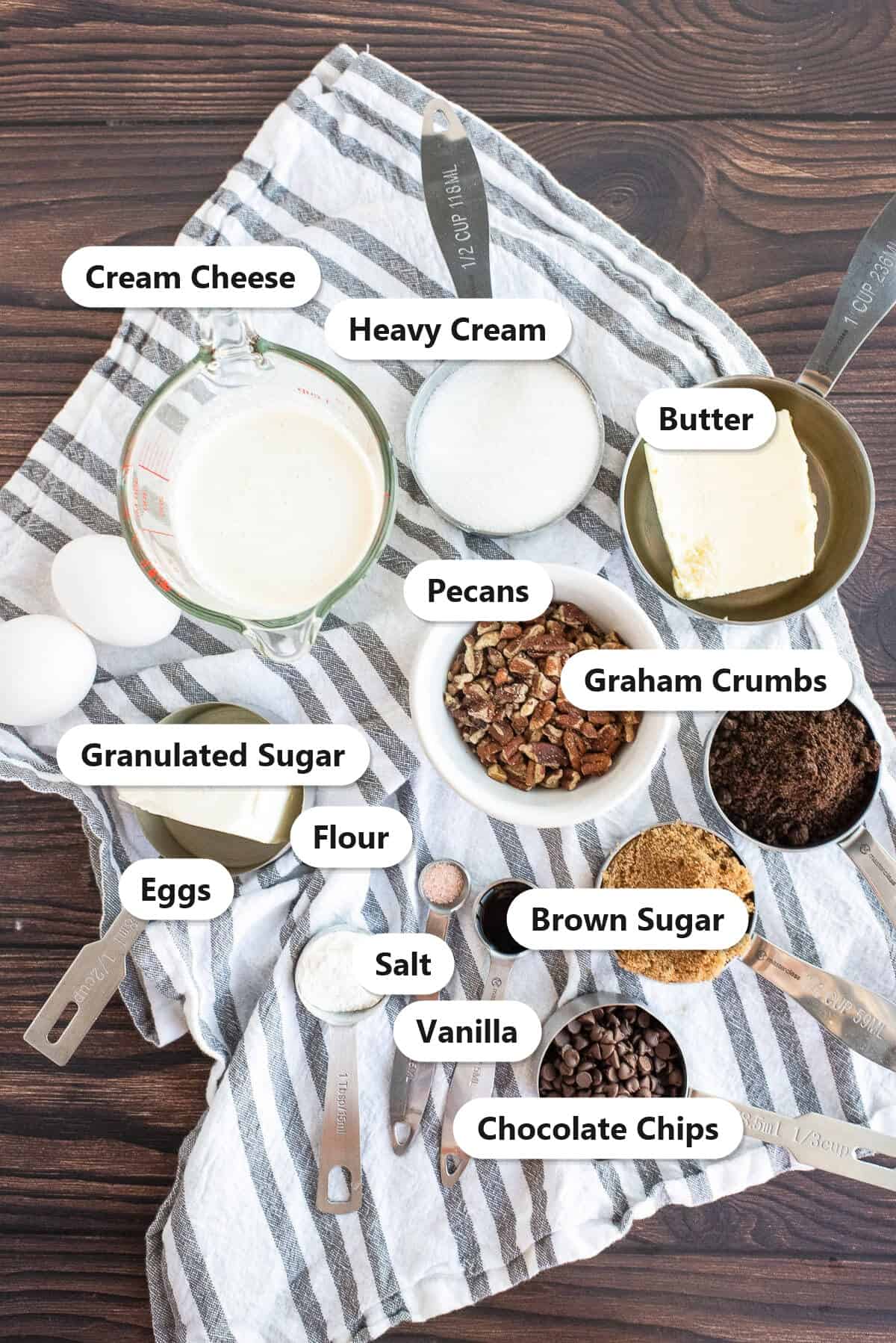 Ingredients to make turtle cheesecake labeled on a striped towel: cream cheese, heavy cream, butter, pecans, graham crumbs, granulated sugar, eggs, flour, salt, vanilla, brown sugar, and chocolate chips.