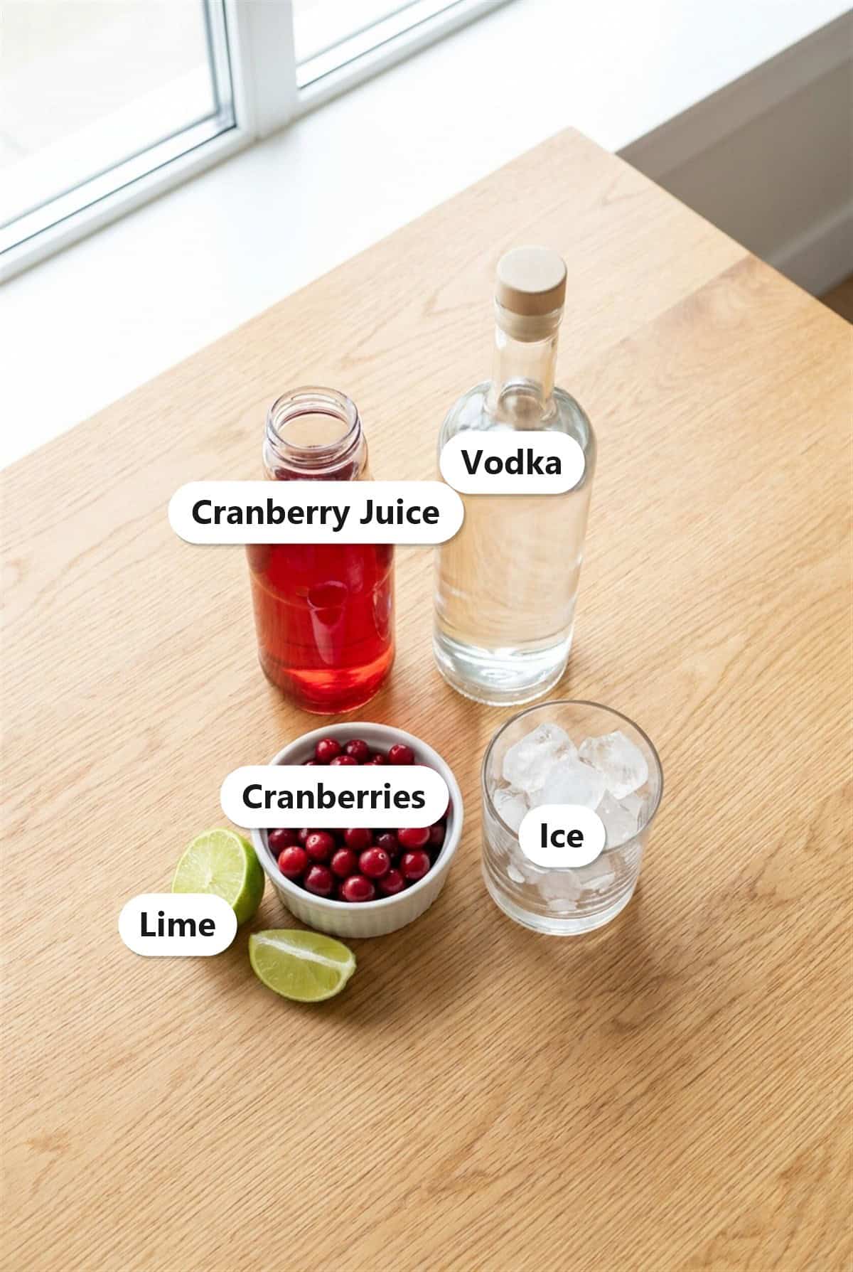 Vodka Cranberry ingredients including vodka, cranberry juice, fresh cranberries, lime, and ice labeled on a wood table.