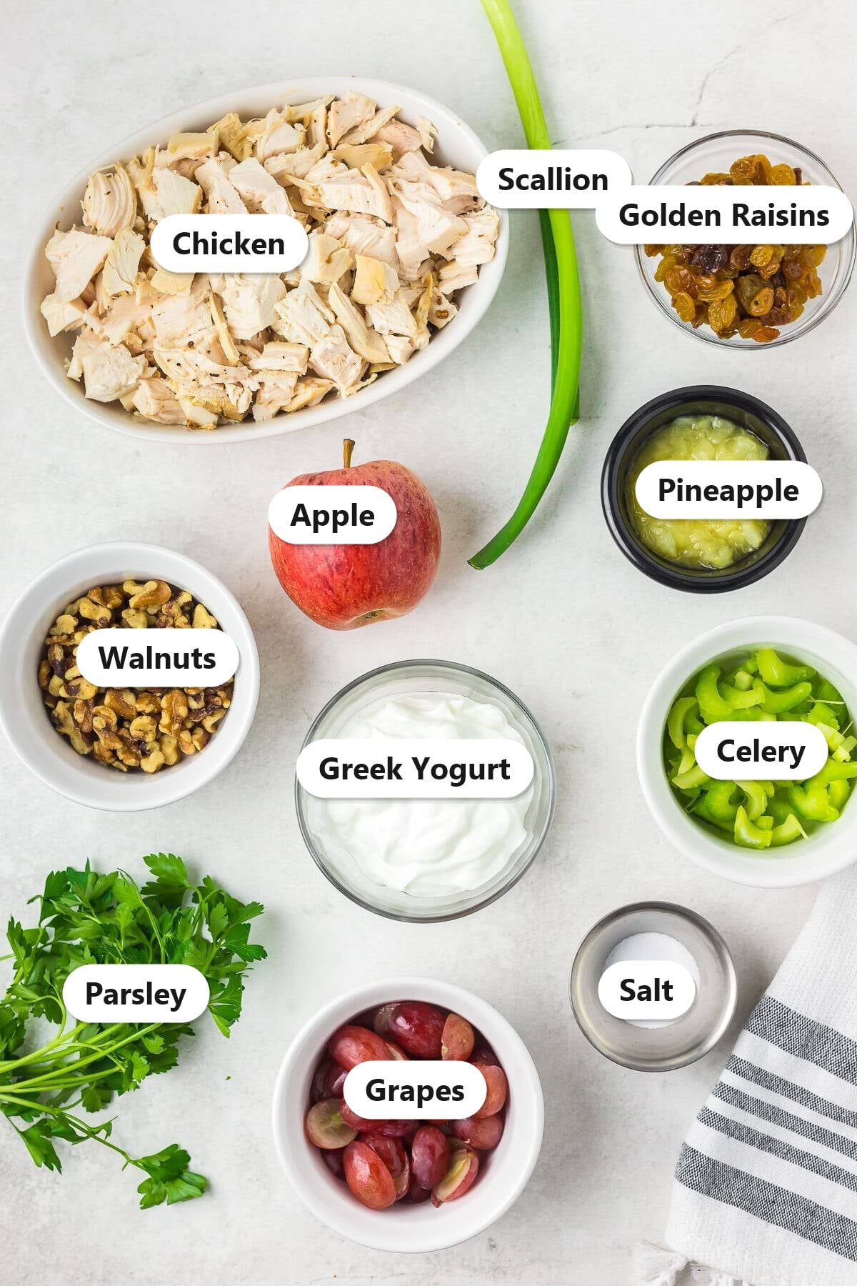 Labeled ingredients to make waldorf chicken salad including chicken, apple, red grapes, celery, walnuts, golden raisins, scallion, parsley, Greek yogurt, crushed pineapple, and salt.