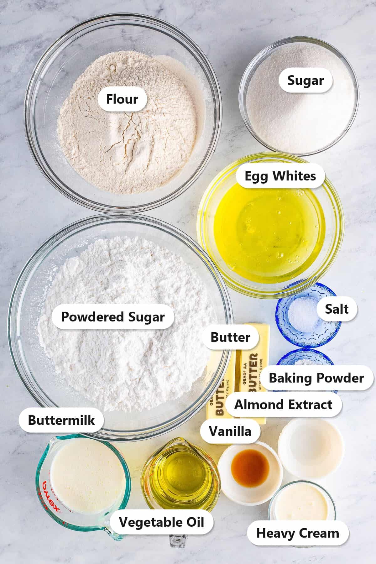 Labeled ingredients for white wedding cake cupcakes including flour, sugar, egg whites, oil, buttermilk, butter, powdered sugar, and almond extract.