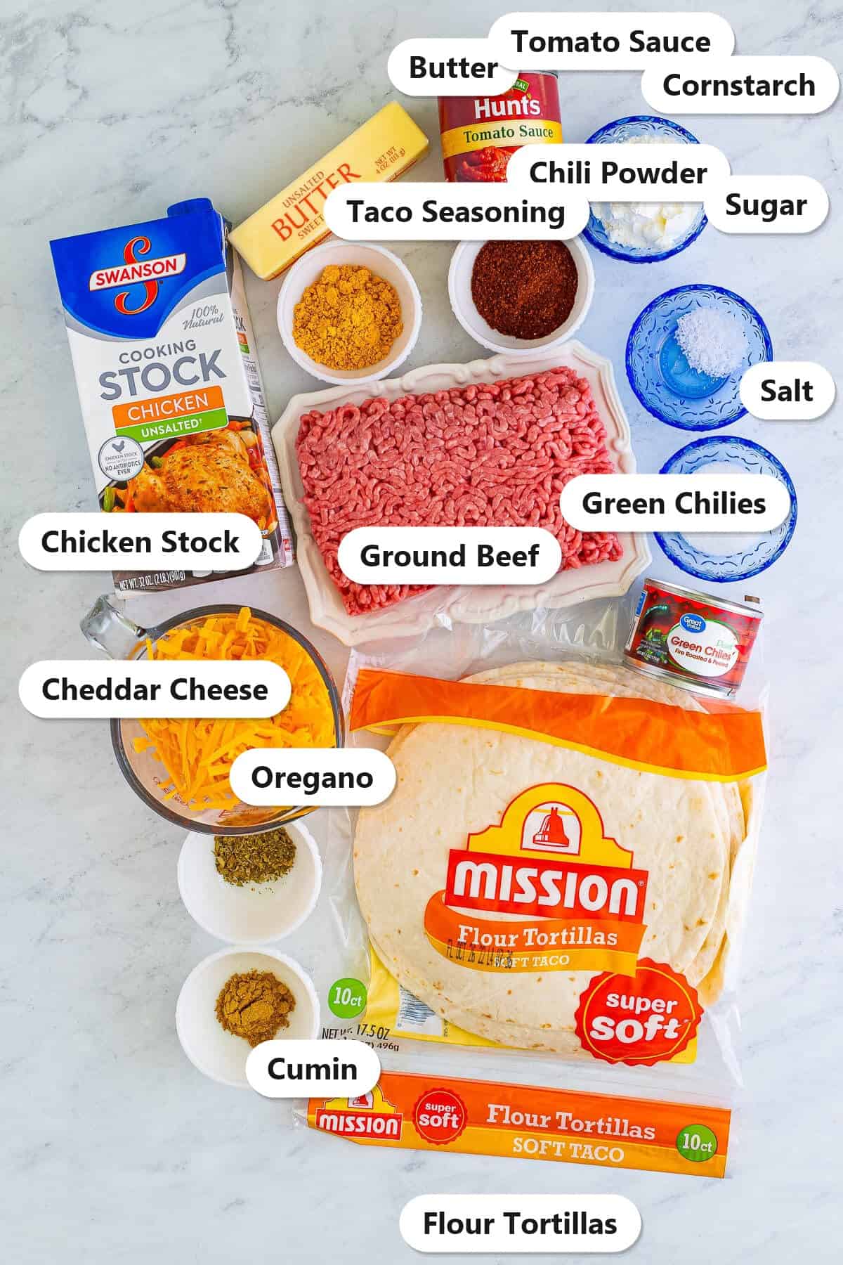 Labeled ingredients to make wet burrito recipe including ground beef, flour tortillas, cheddar cheese, chicken stock, butter, tomato sauce, green chilies, and spices.