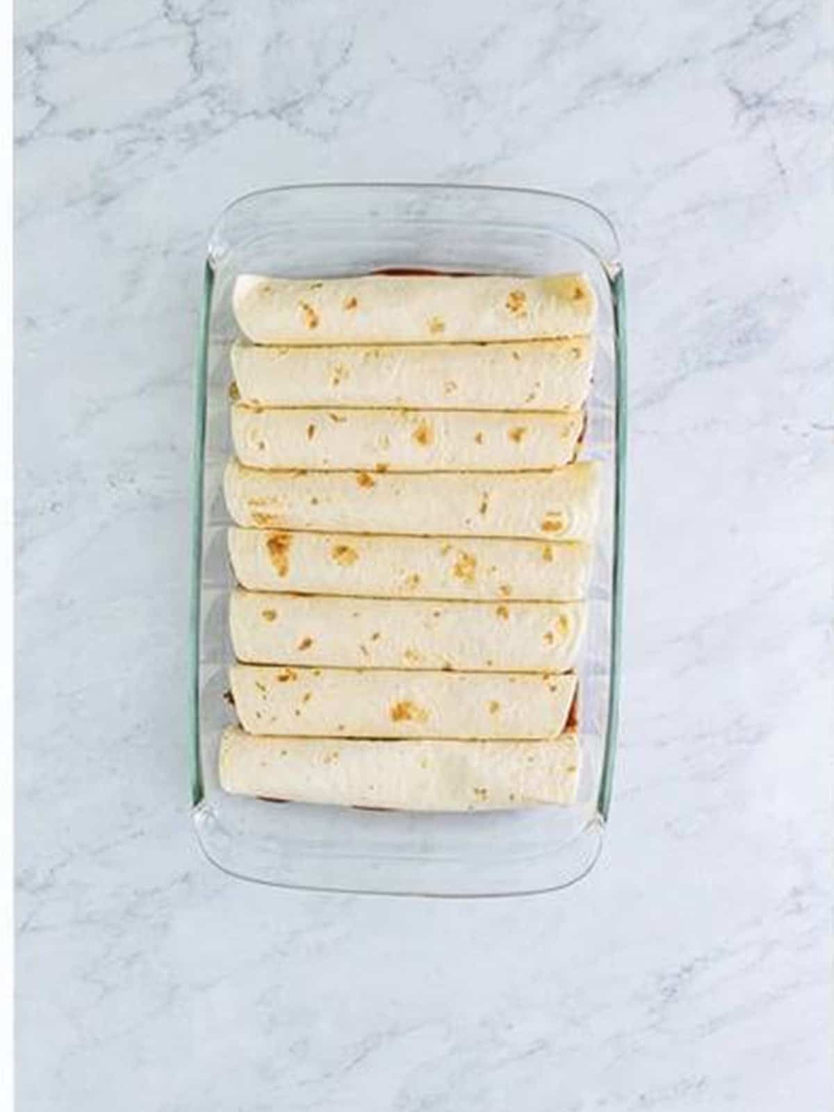 Rolled burritos lined up seam side down in the sauced baking dish.