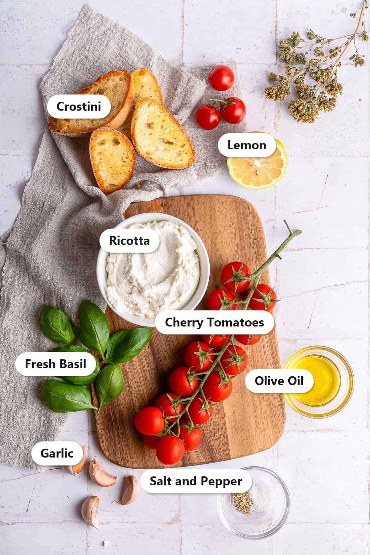 Labeled ingredients for whipped ricotta dip including ricotta, cherry tomatoes, garlic, olive oil, fresh basil, lemon, and crostini.