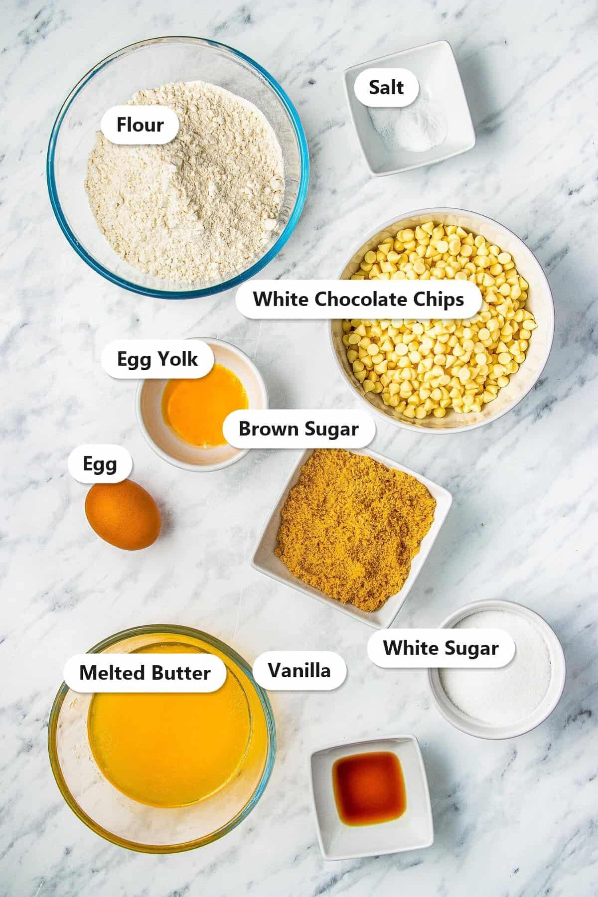 Labeled ingredients to make white chocolate chip cookies including flour, white chocolate chips, brown sugar, white sugar, melted butter, egg, and vanilla.