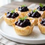 A white plate of wild blueberry no bake cheesecake bites in phyllo cups topped with blueberry sauce and fresh mint.
