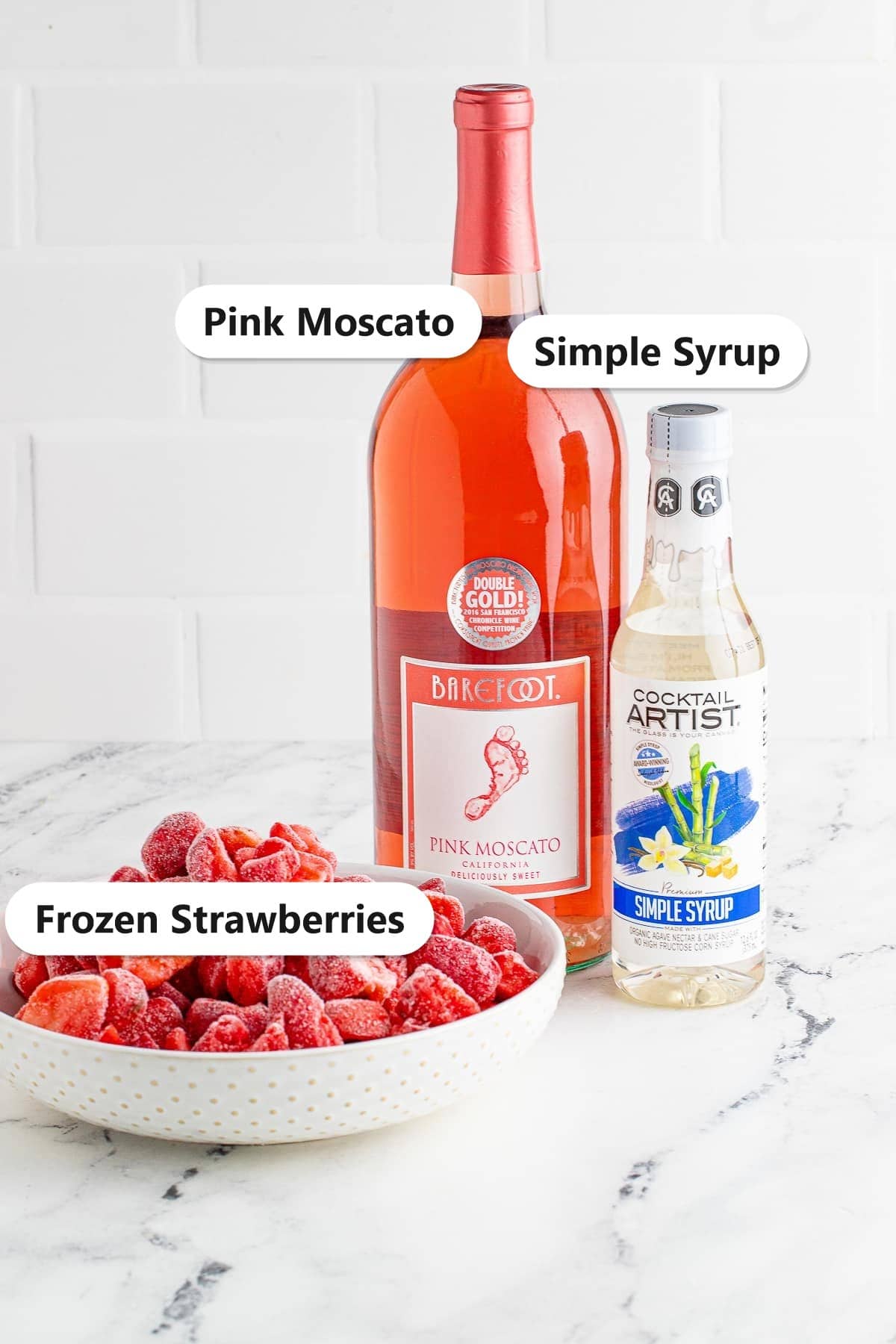 Labeled ingredients to make a wine slushie including pink Moscato, simple syrup, and frozen strawberries.