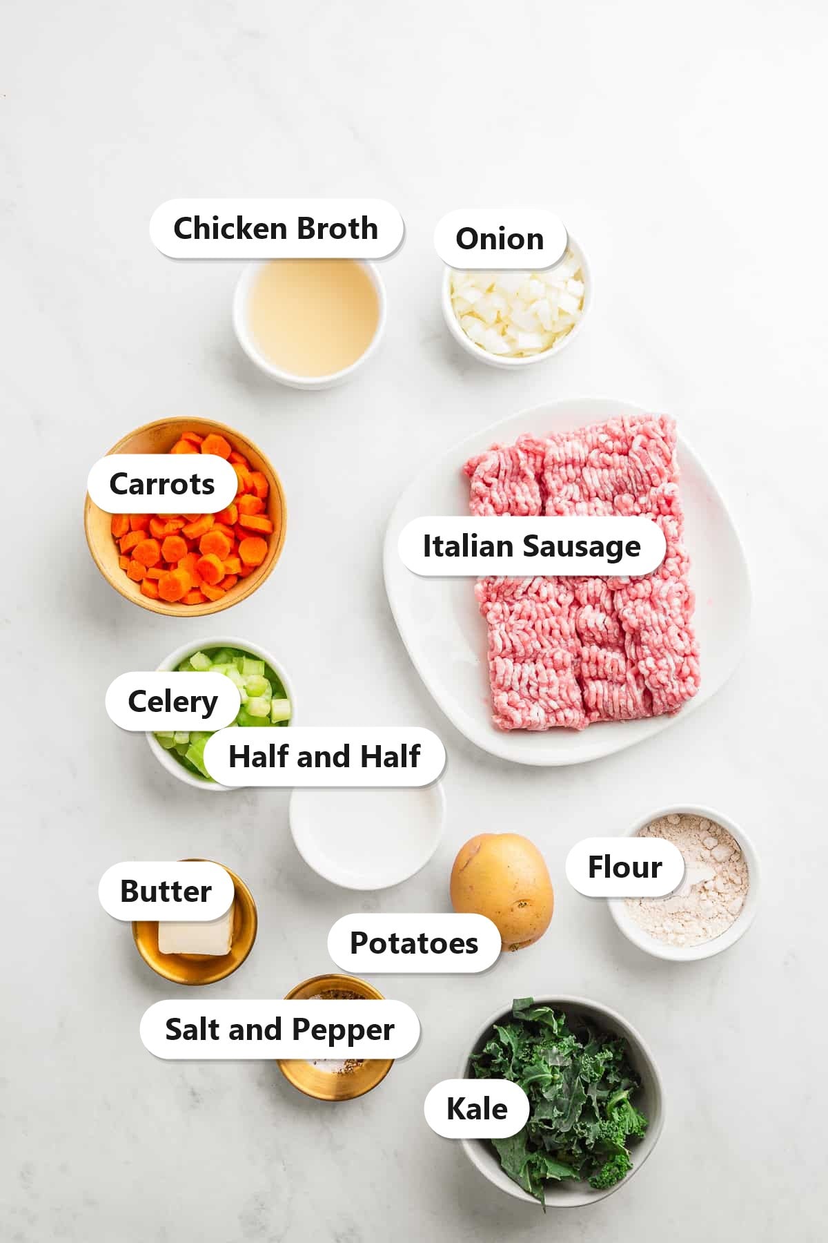 Zuppa Toscana ingredients including Italian sausage, kale, golden potatoes, carrots, celery, onion, broth, flour, butter, and half and half labeled on a marble surface.