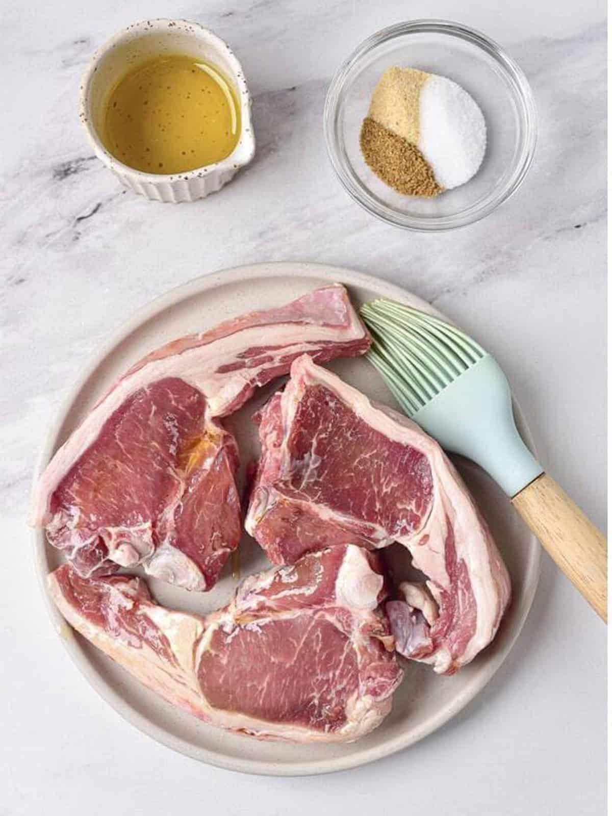 Raw lamb chops on a plate being brushed with olive oil.