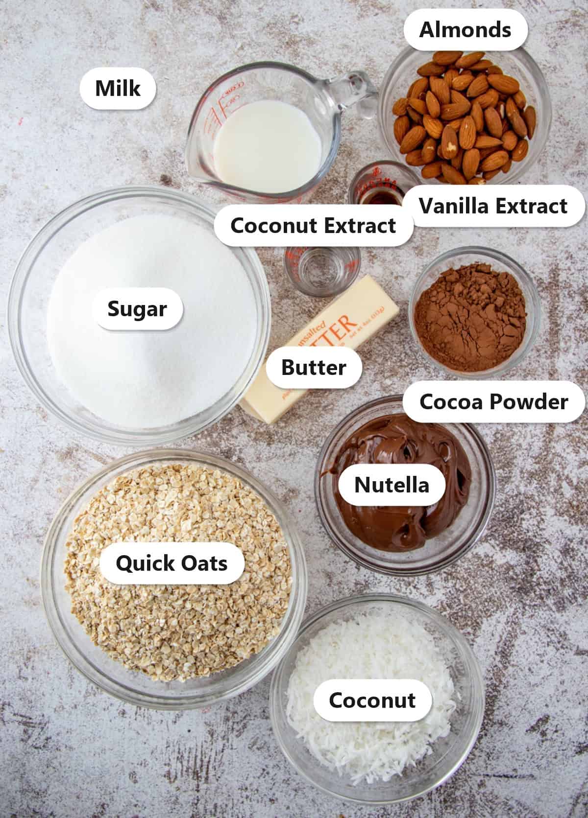 Labeled ingredients to make almond joy cookies including oats, coconut, cocoa powder, Nutella, almonds, butter, sugar, and milk