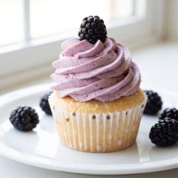 An angel food cupcake topped with a swirl of pale purple blackberry buttercream and a fresh blackberry.