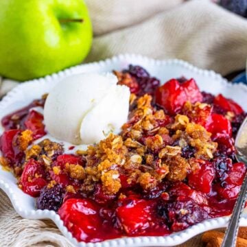 A serving of apple and blackberry crumble topped with a scoop of vanilla ice cream in a white bowl.