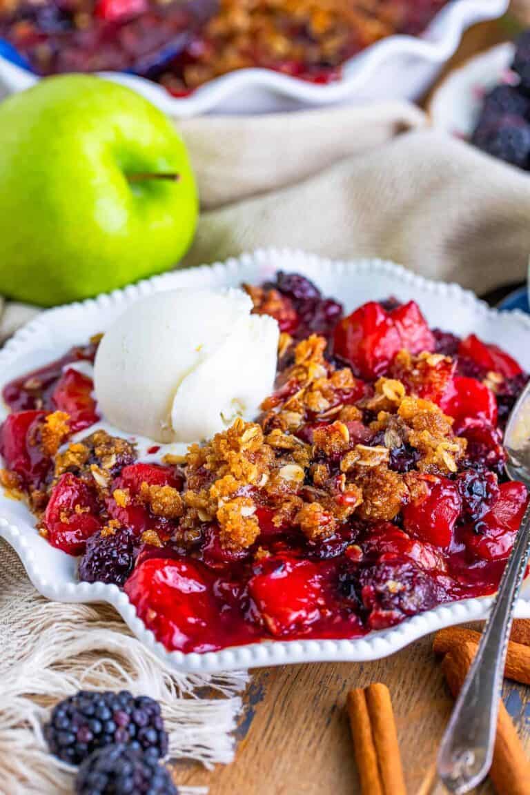A serving of apple and blackberry crumble topped with a scoop of vanilla ice cream in a white bowl.