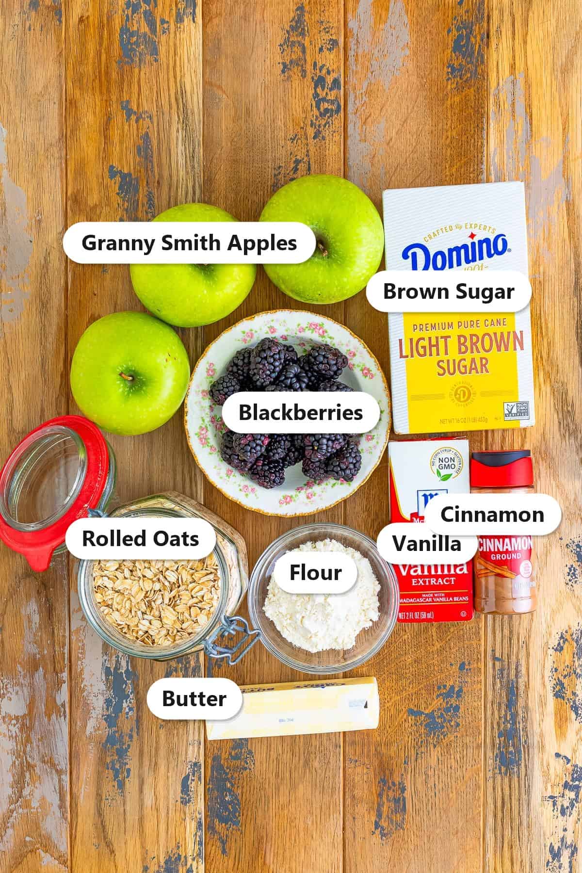 Labeled ingredients to make apple and blackberry crumble including Granny Smith apples, blackberries, brown sugar, rolled oats, flour, butter, vanilla, and cinnamon.