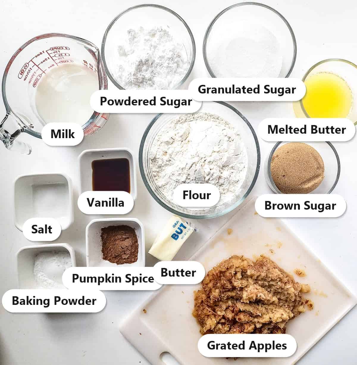 Labeled ingredients to make Apple Bread: milk, powdered sugar, granulated sugar, melted butter, flour, brown sugar, vanilla, salt, pumpkin spice, baking powder, butter, and grated apples.