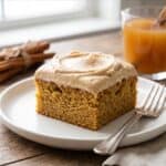 A frosted square slice of Apple Cider Cake on a white plate with a glass of apple cider.