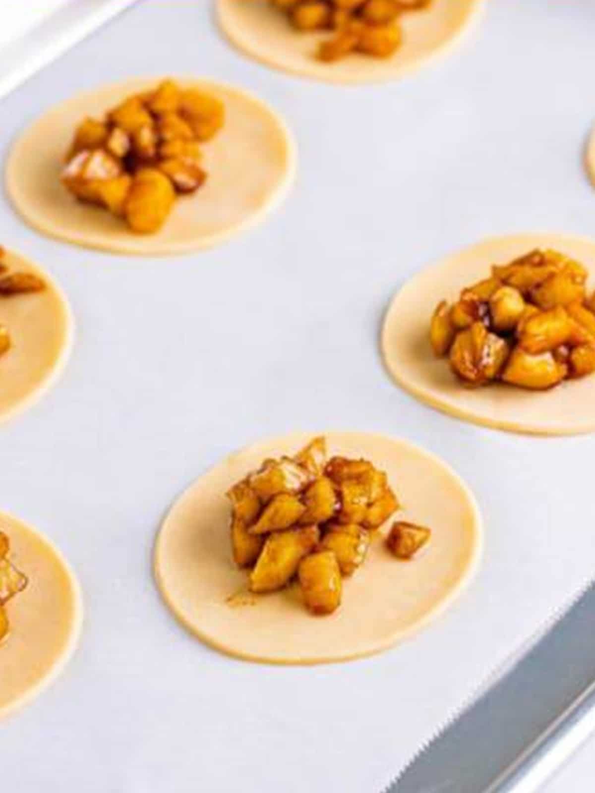 Apple pie filling spooned onto pie crust circles on a parchment lined tray.