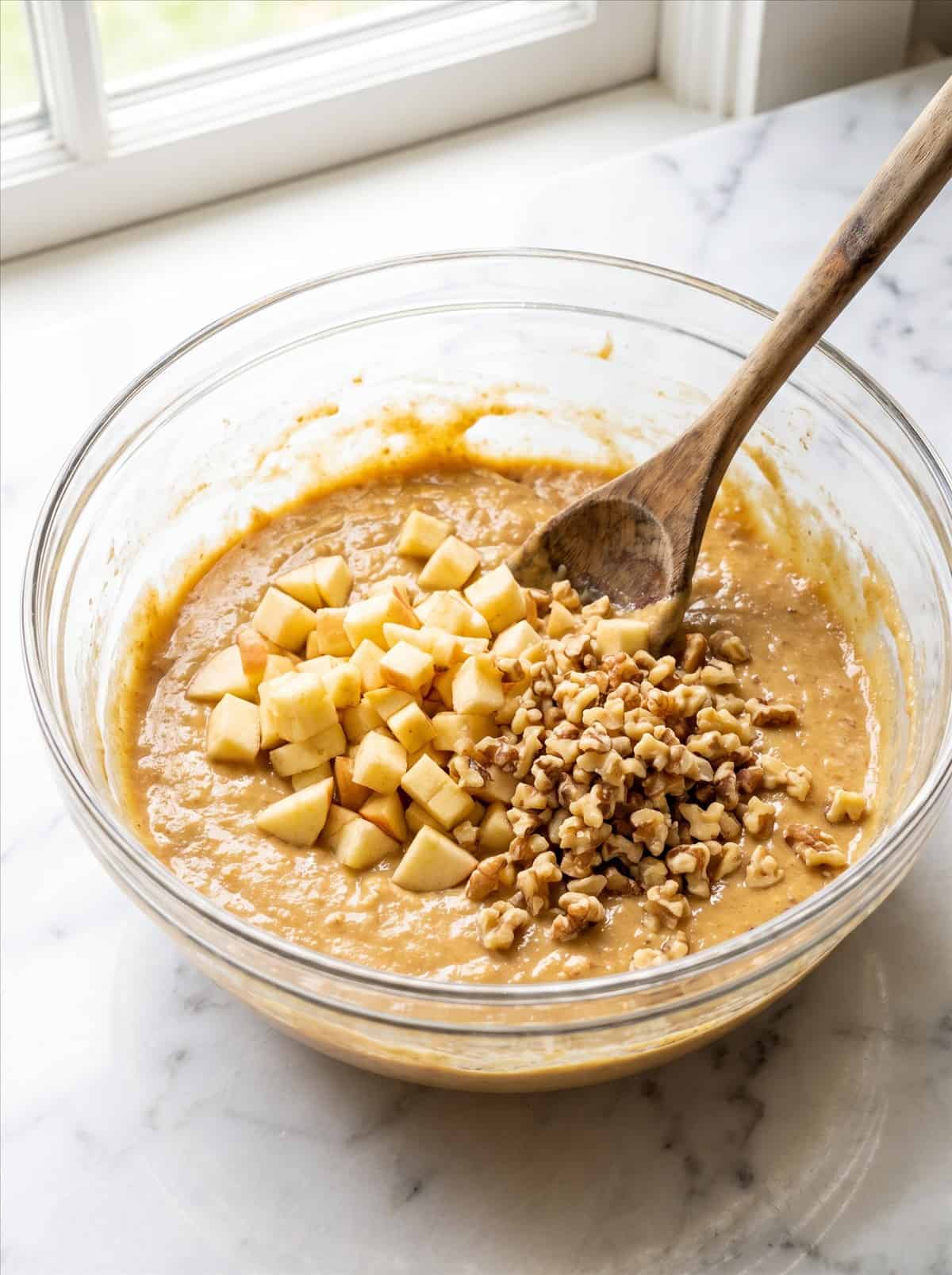 Chopped apples and walnuts on top of thick golden batter in a glass mixing bowl ready to be folded in for apple walnut bread.