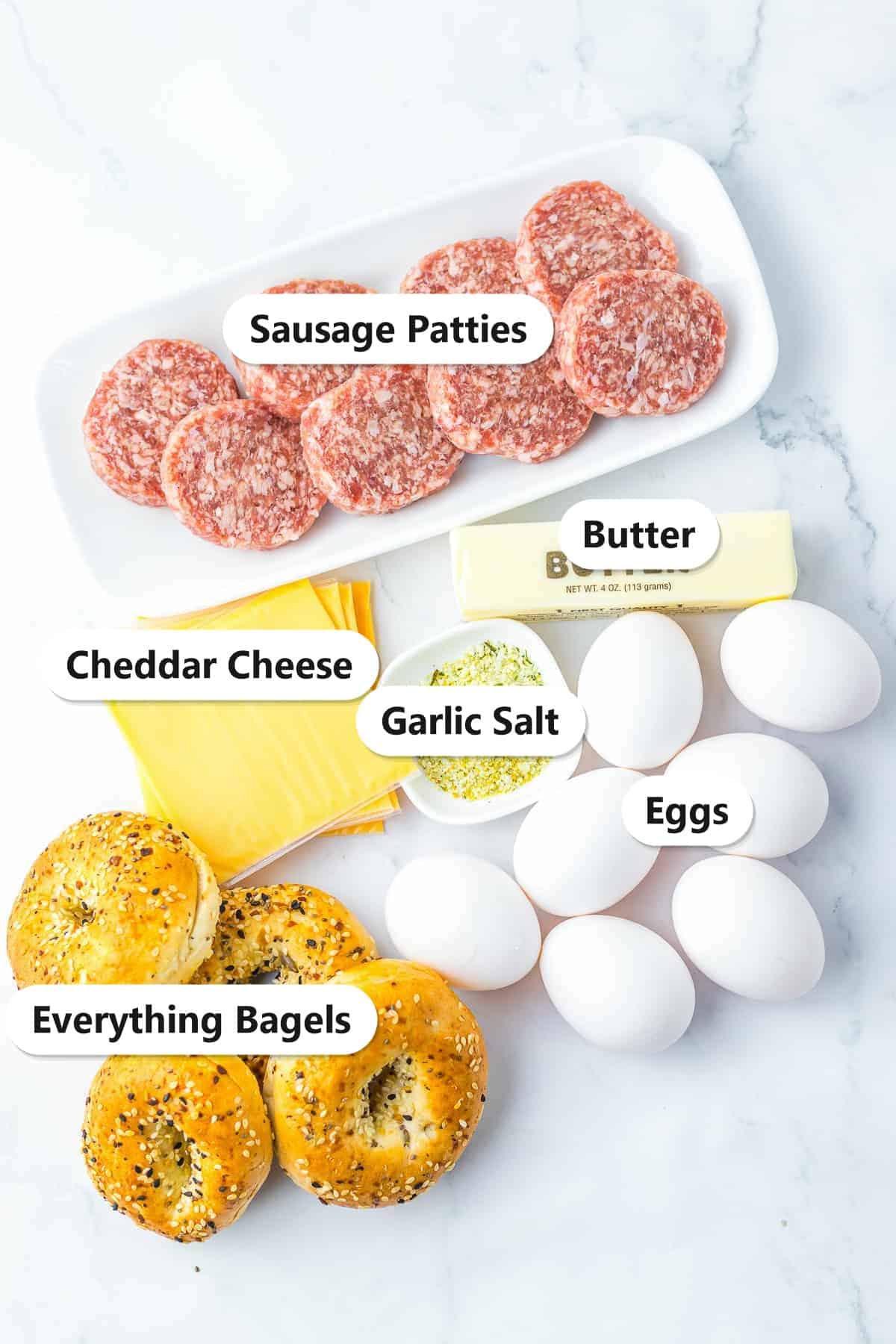 Labeled ingredients to make bagel breakfast sliders including sausage patties, eggs, everything bagels, cheddar cheese, butter, and garlic salt.