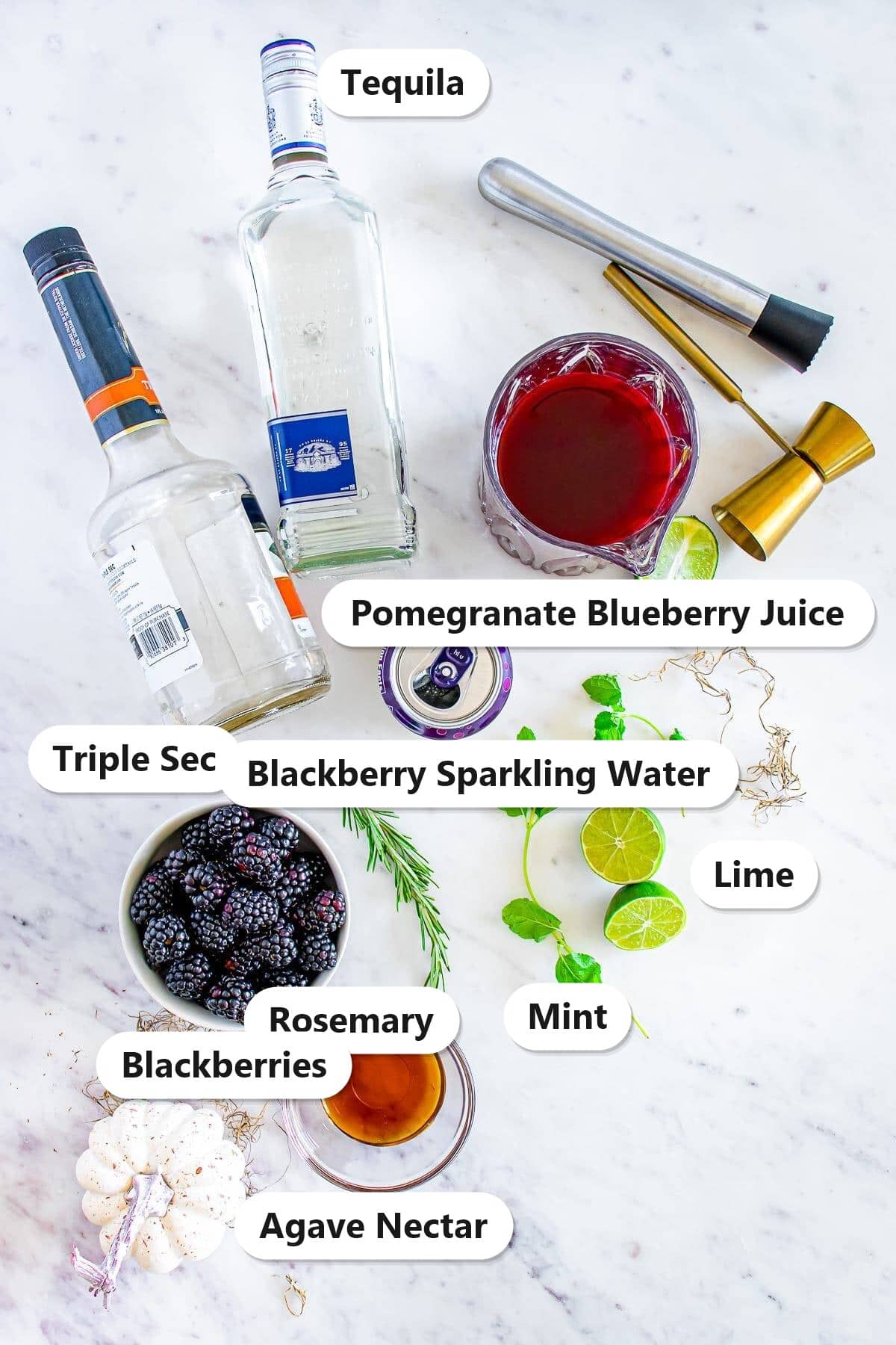 Ingredients to make a blackberry margarita labeled on a marble surface: tequila, triple sec, pomegranate blueberry juice, lime, blackberry sparkling water, mint, rosemary, blackberries, and agave nectar.