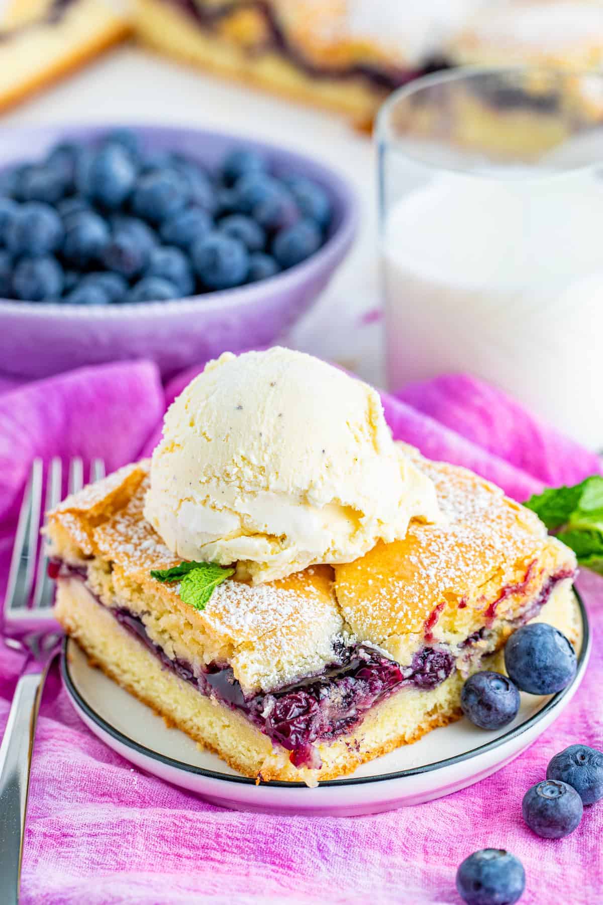 A blueberry bar topped with a scoop of vanilla ice cream on a plate with fresh blueberries.