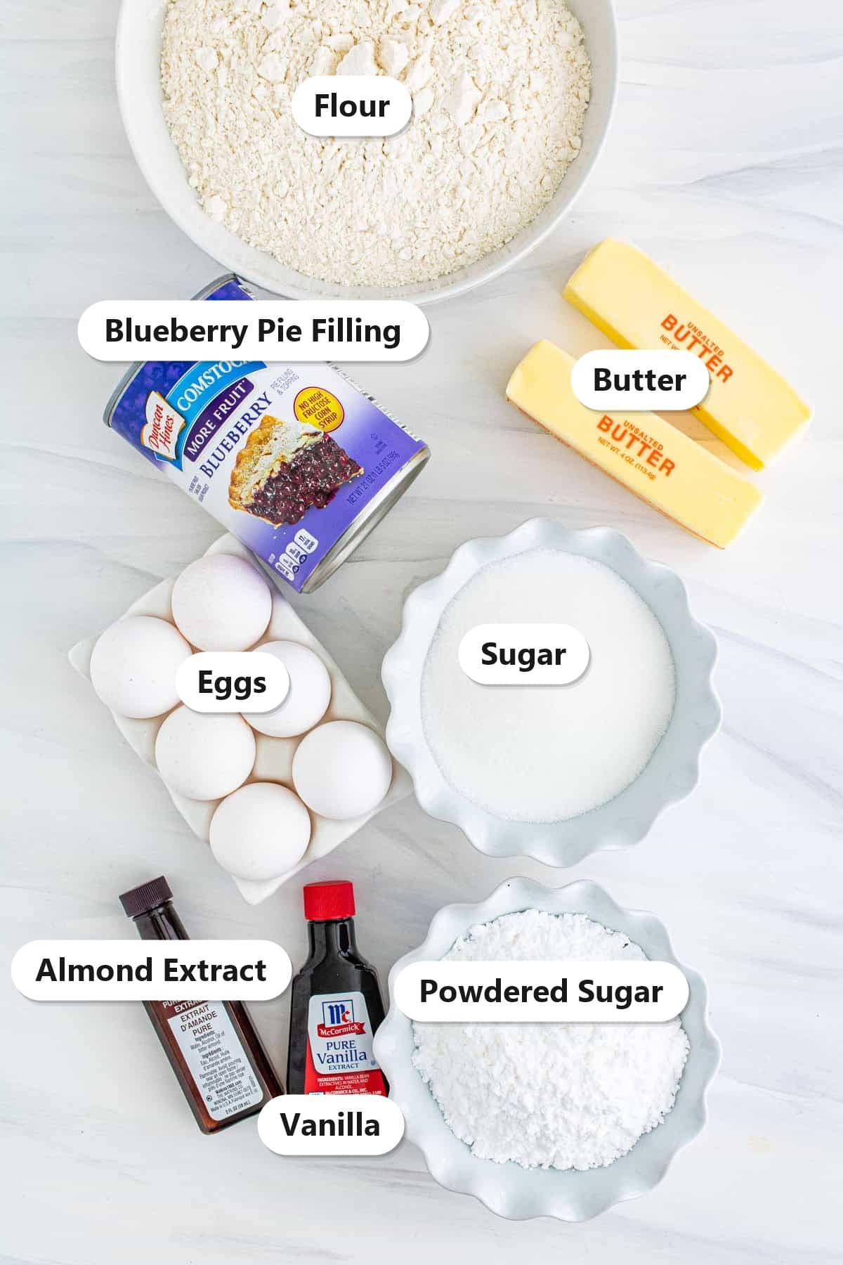 Labeled ingredients to make blueberry bars including flour, butter, sugar, eggs, vanilla, almond extract, blueberry pie filling, and powdered sugar.