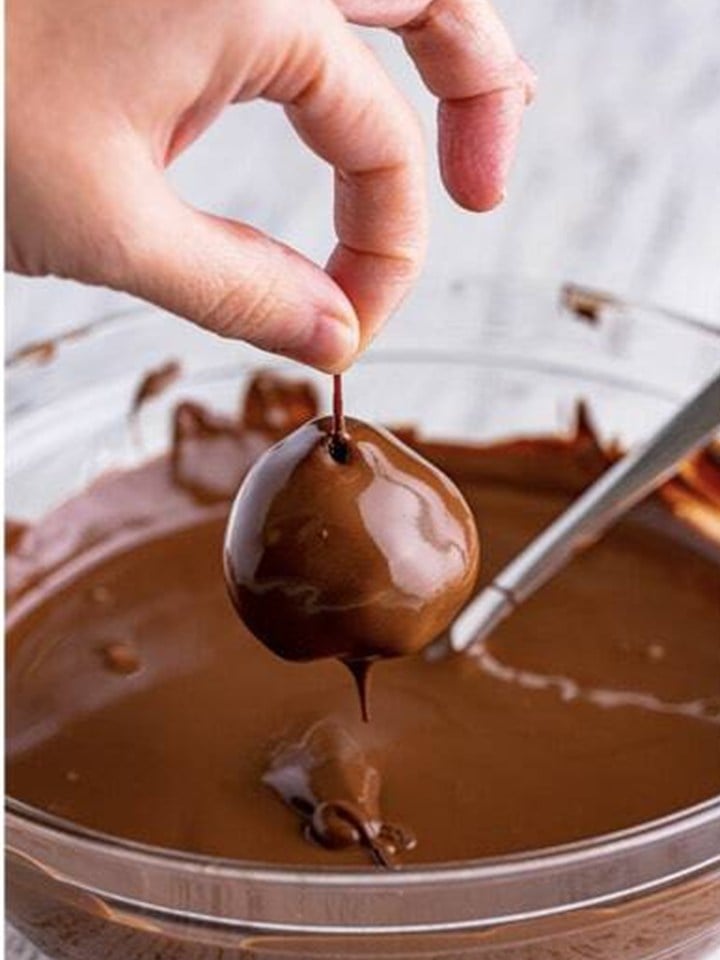 A brownie truffle held by the cherry stem being dipped into melted chocolate coating.