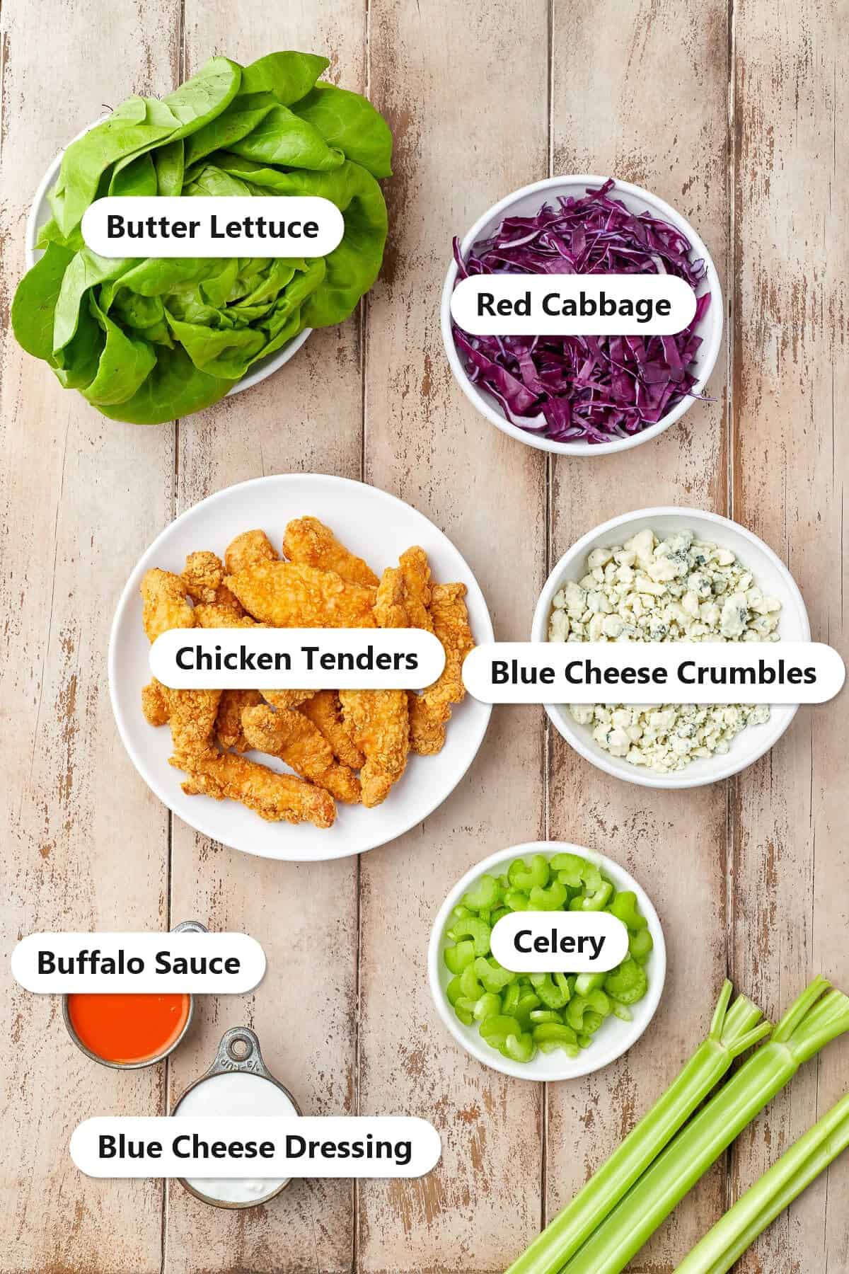 Labeled ingredients to make buffalo chicken lettuce wraps including chicken tenders, buffalo sauce, butter lettuce, red cabbage, celery, blue cheese crumbles, and blue cheese dressing.