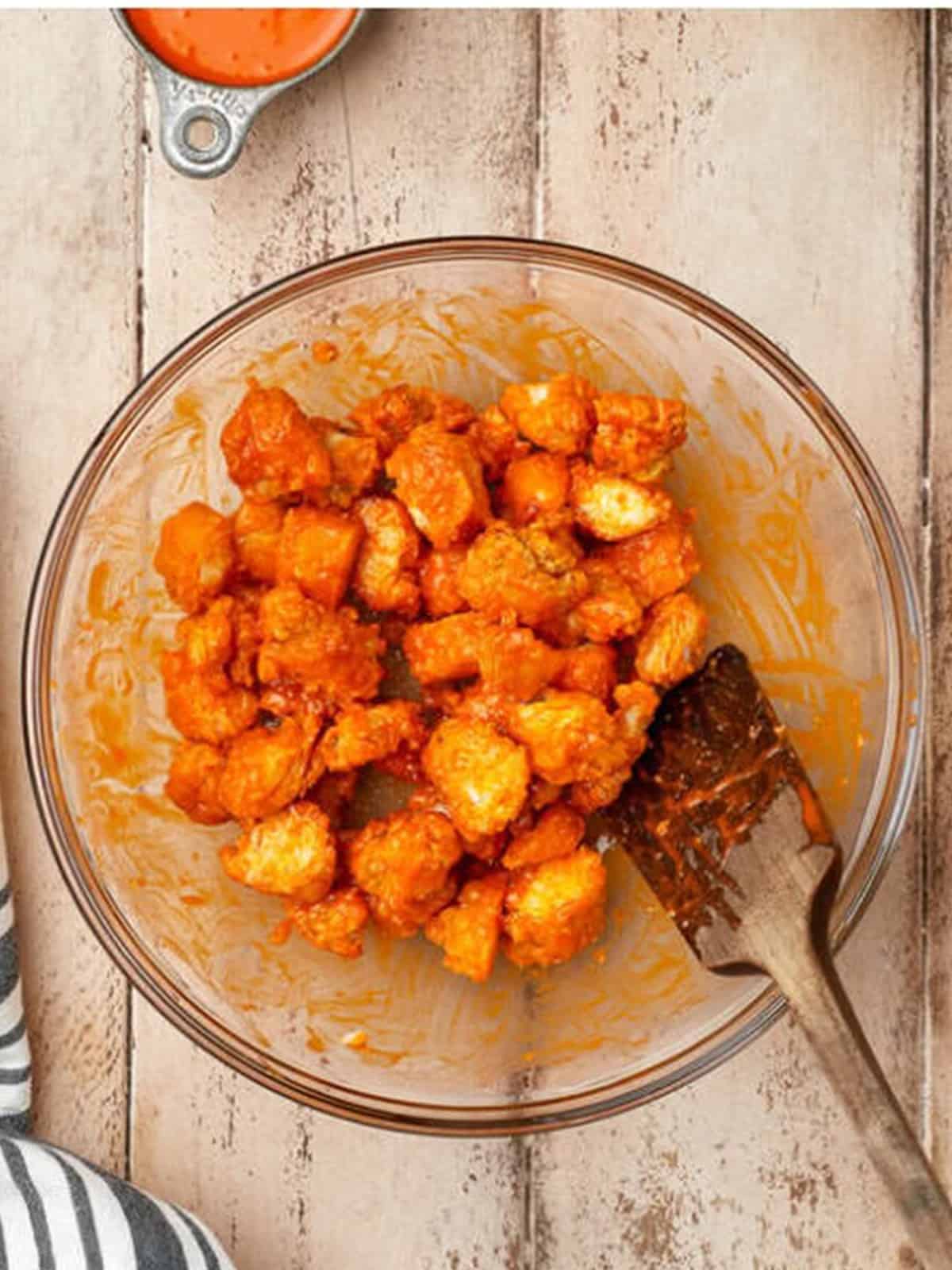 Bite-sized chicken pieces tossed with buffalo sauce in a glass bowl until fully coated.