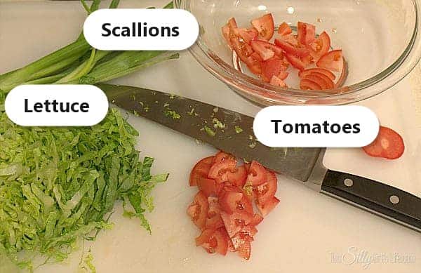 Shredded lettuce, chopped tomatoes, and scallions labeled on a cutting board for chicken pitas.