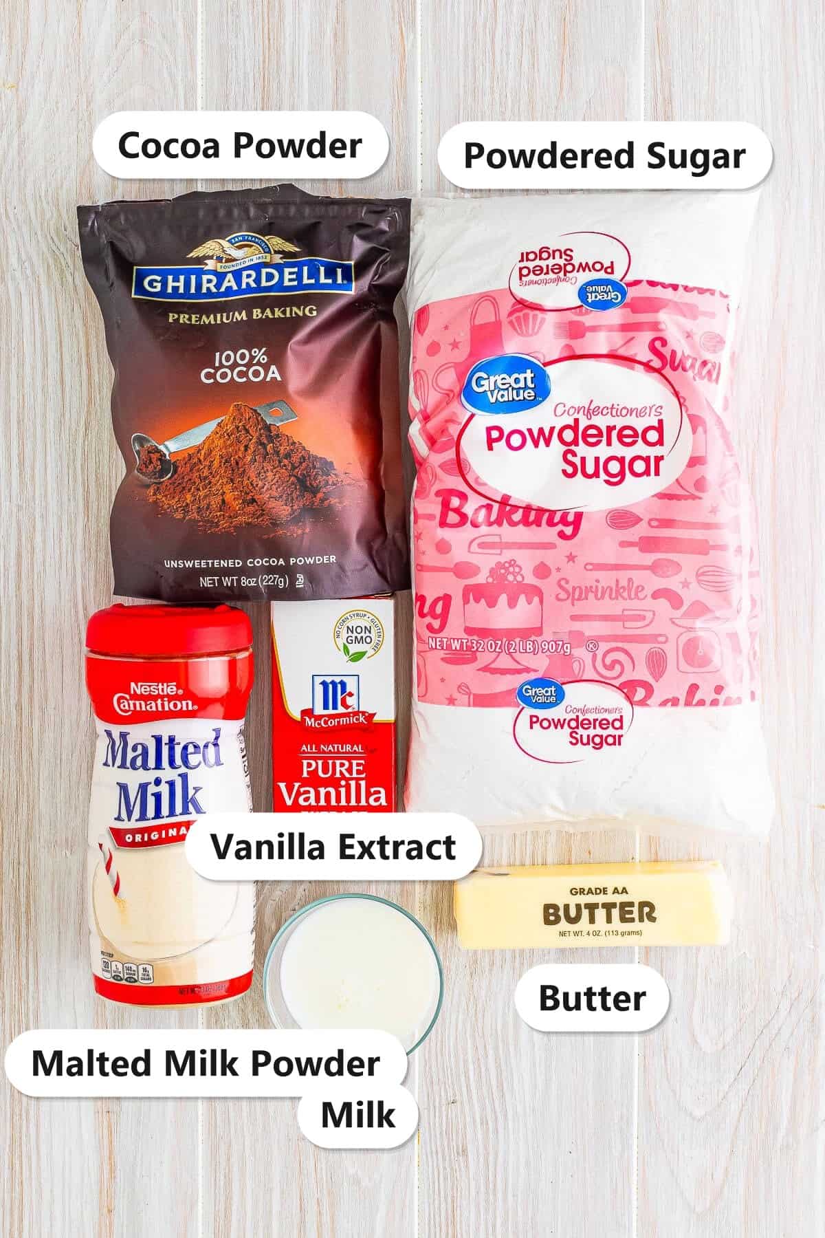Ingredients to make chocolate buttercream frosting labeled on a wood surface: cocoa powder, powdered sugar, malted milk powder, vanilla extract, butter, and milk.