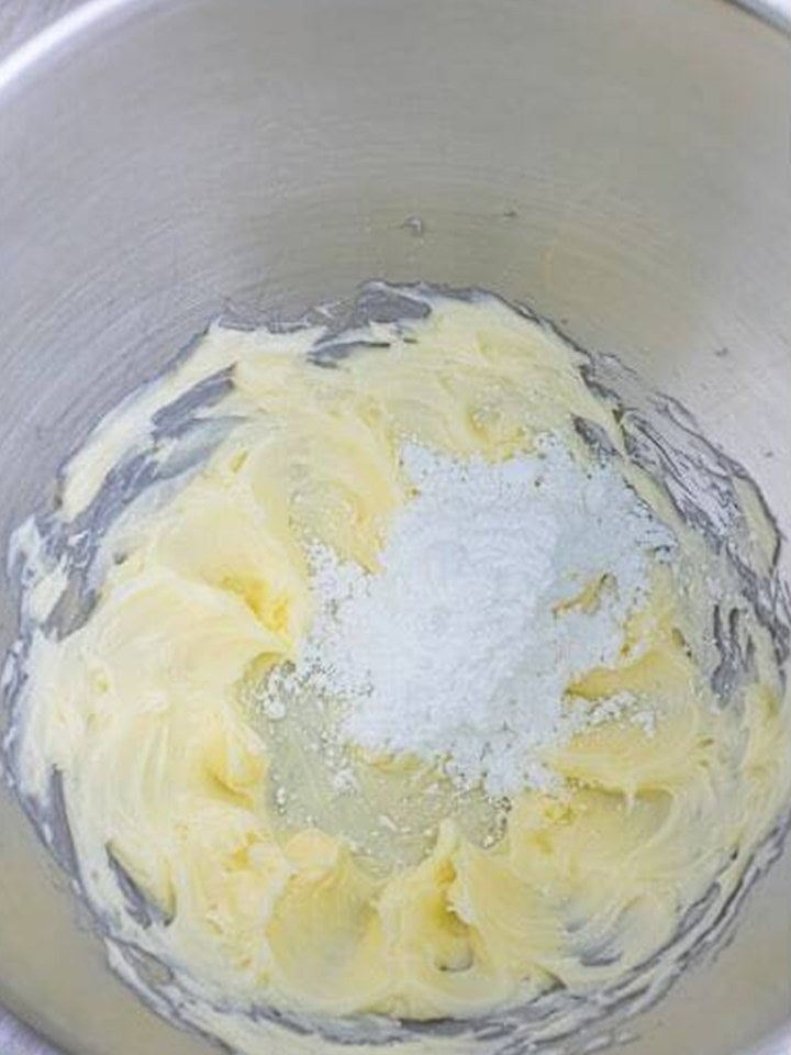 Softened butter whipped in a stand mixer bowl with powdered sugar being added for chocolate buttercream frosting.