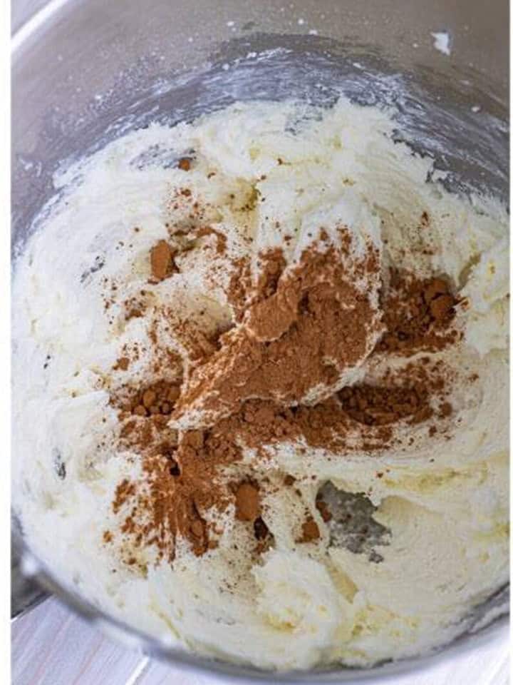 Cocoa powder added on top of the whipped butter mixture in a stand mixer bowl.