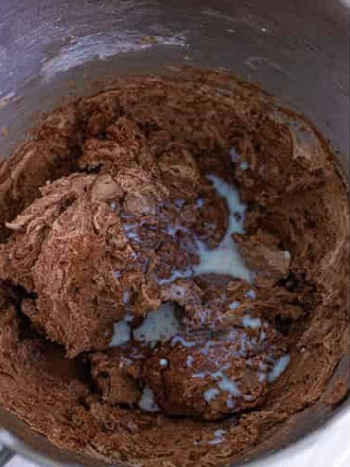 Milk being mixed into the chocolate frosting base in a stand mixer bowl.