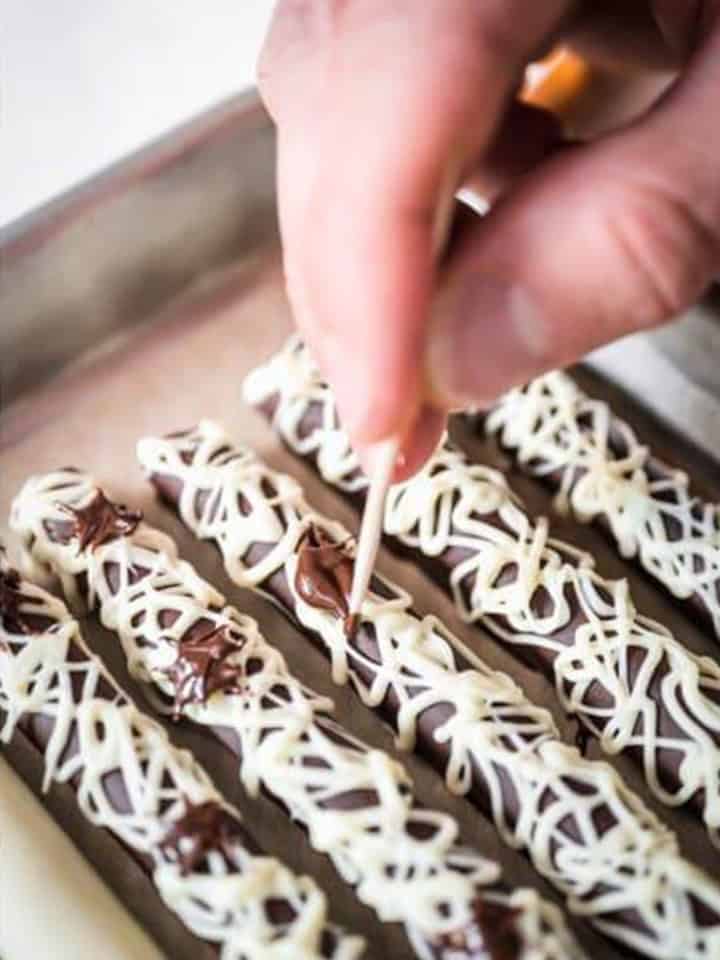 Chocolate covered pretzel rods on a tray being decorated with piped white chocolate designs.