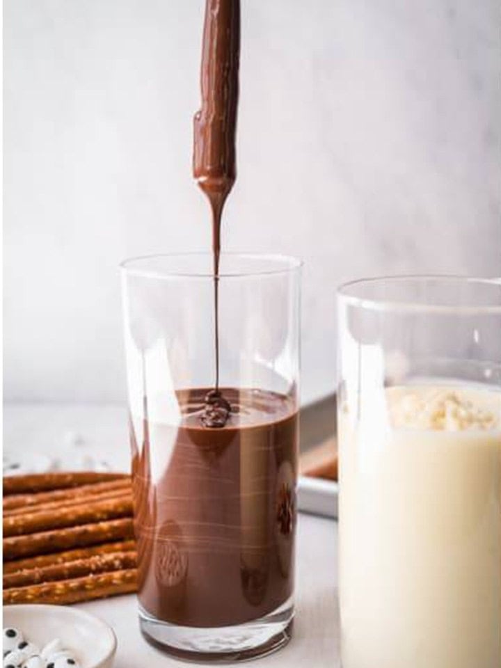 Pretzel rod being dipped into a tall glass of melted semisweet chocolate.