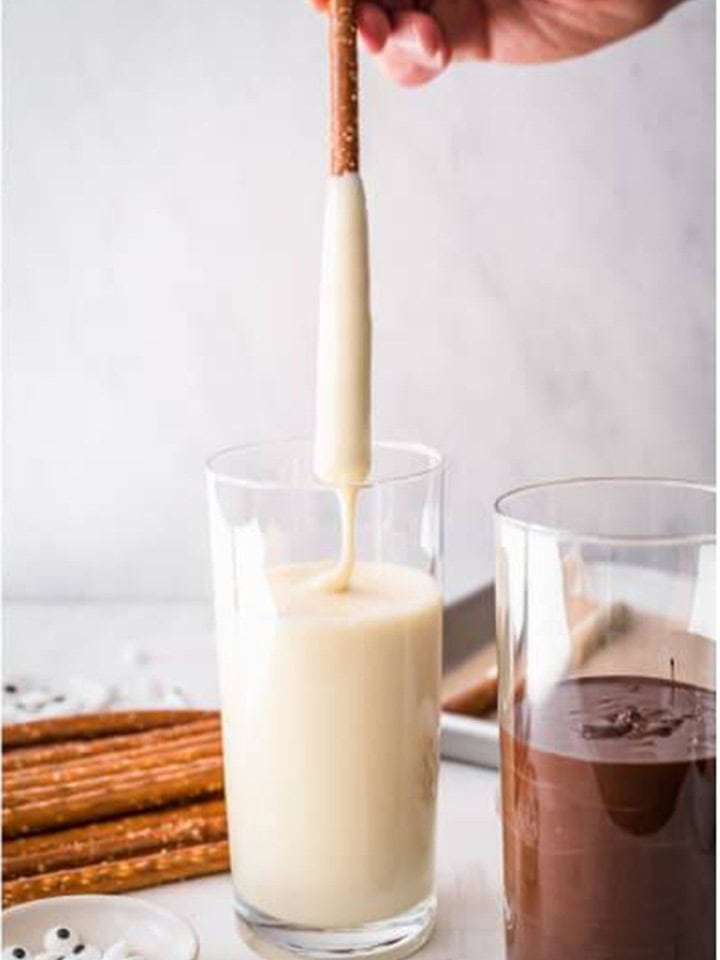 Pretzel rod being dipped three quarters deep into a tall glass of melted white chocolate.
